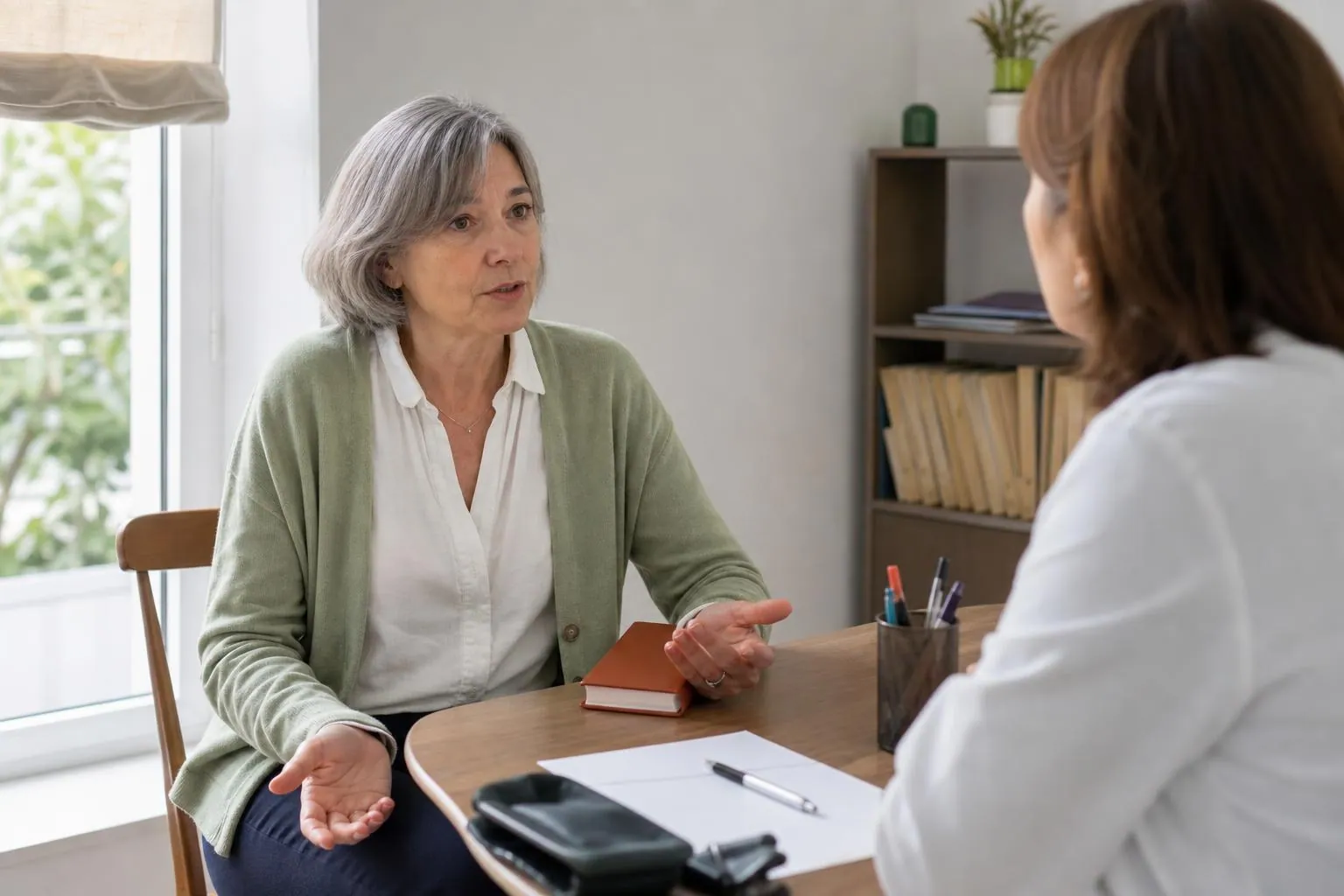 Femme d’une cinquantaine d’années assise face à une médecin dans un cabinet clair, une ordonnance papier et un stylo posés sur le bureau en bois